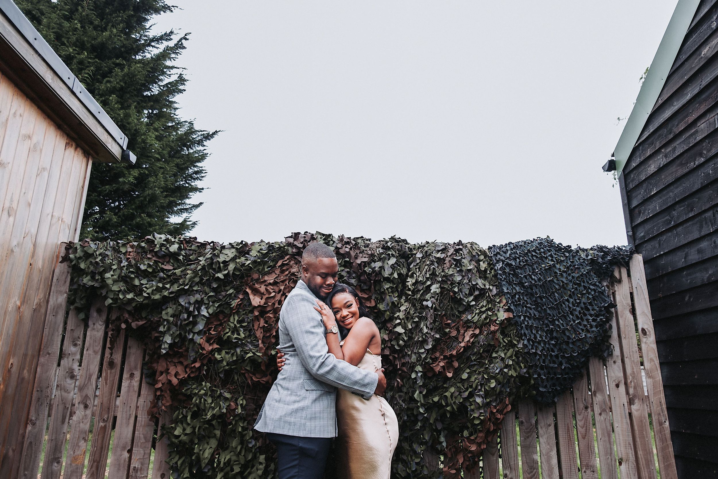 A couple embraces in front of a rustic wooden wall covered with climbing ivy and greenery.