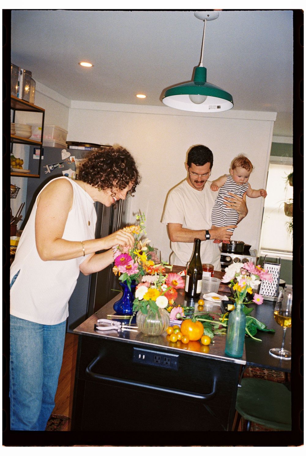 Dad is holding baby in the kitchen while cooking dinner, Mom cuts fresh flowers at the counter on flash 35mm film in Biddeford Maine