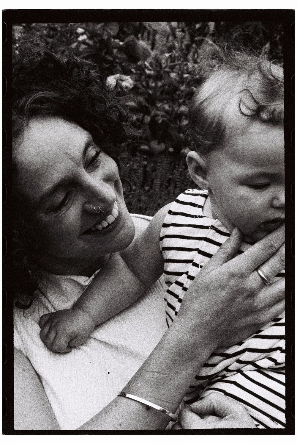 Mother holds her daughter up in the air and smiles sweetly while her baby girl wears a striped outfit in front of their home garden in Maine