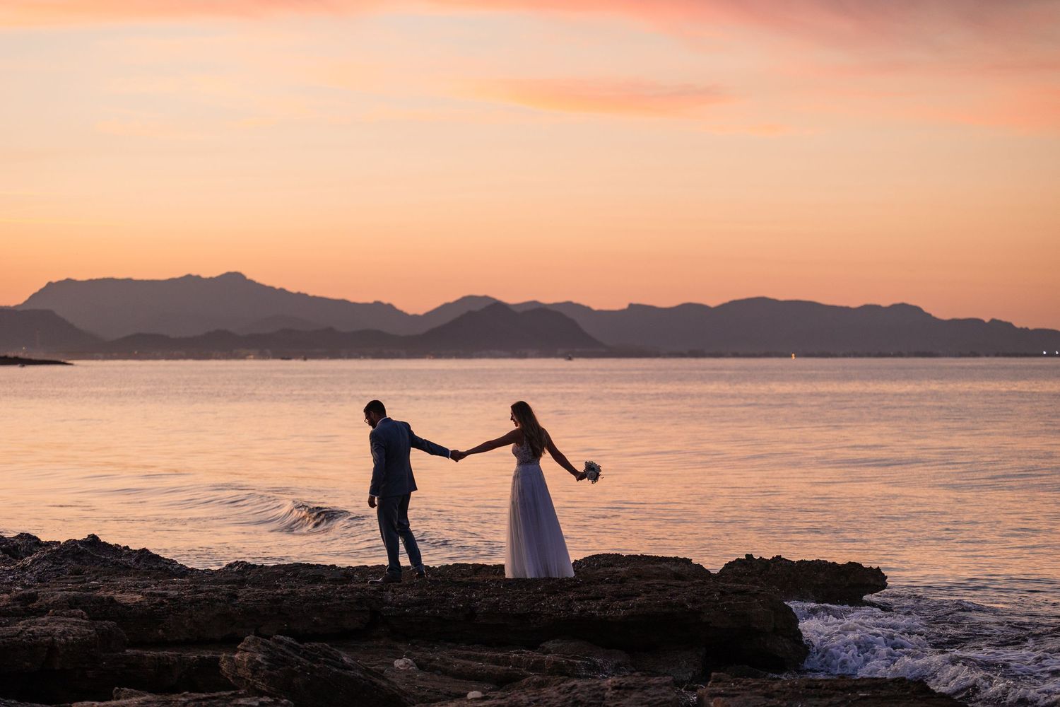 Silhouette eines Hochzeitspaares, das sich an einer felsigen Küste auf Mallorca bei rosa Sonnenuntergang am Meer an den Händen hält.