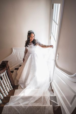 Beautiful bride wearing her dress thoughtfully stares out window as she stands on staircase