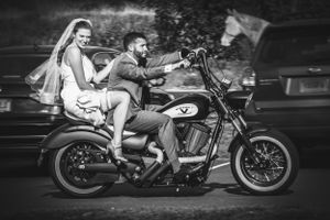 Bride in wedding dress is driven by groom on the back of motor cycle