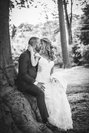 Bride sits on groom's lap as they kiss, leaning on a large tree