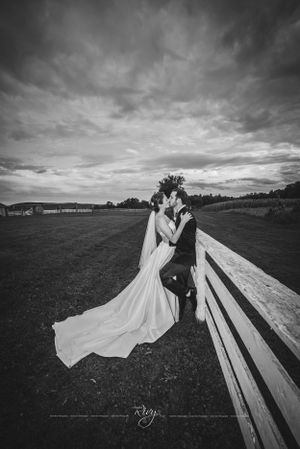 Dramatic black and white of majestic sky in back as bride and groom lean on fence and kiss