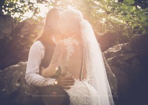 Groom holds his bride closely under tree canopy with sun bursting through