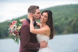 Elegant bride is kissed by handsome groom in red jacket near a lake