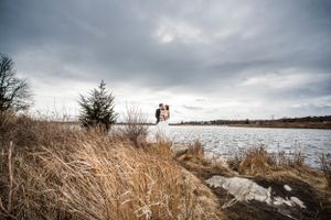 Bride and groom stand near large lake, surrounded by tall grass and fir trees