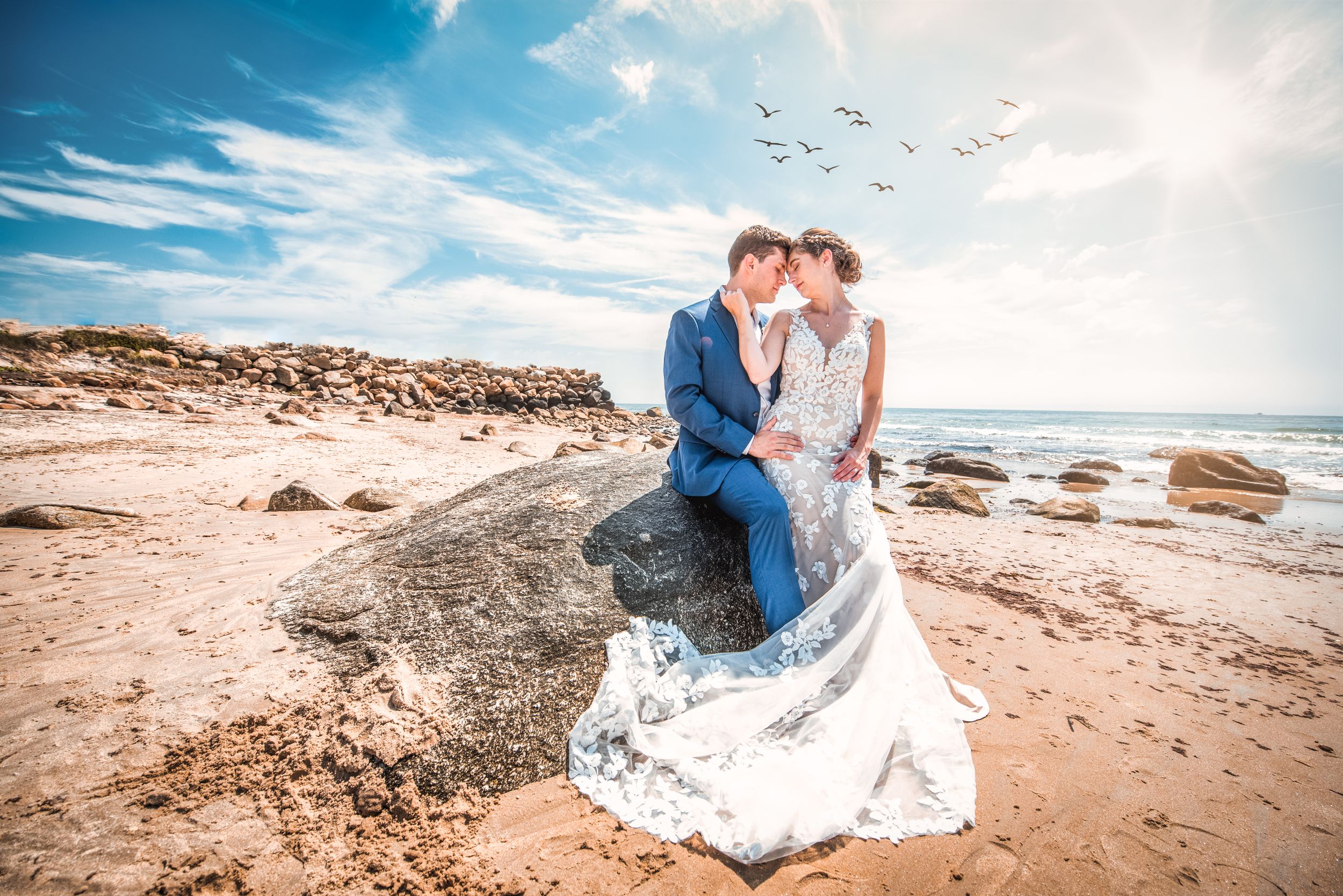 Groom holds bride as they sit elegantly on a rock at the beach with ocean and sun in back