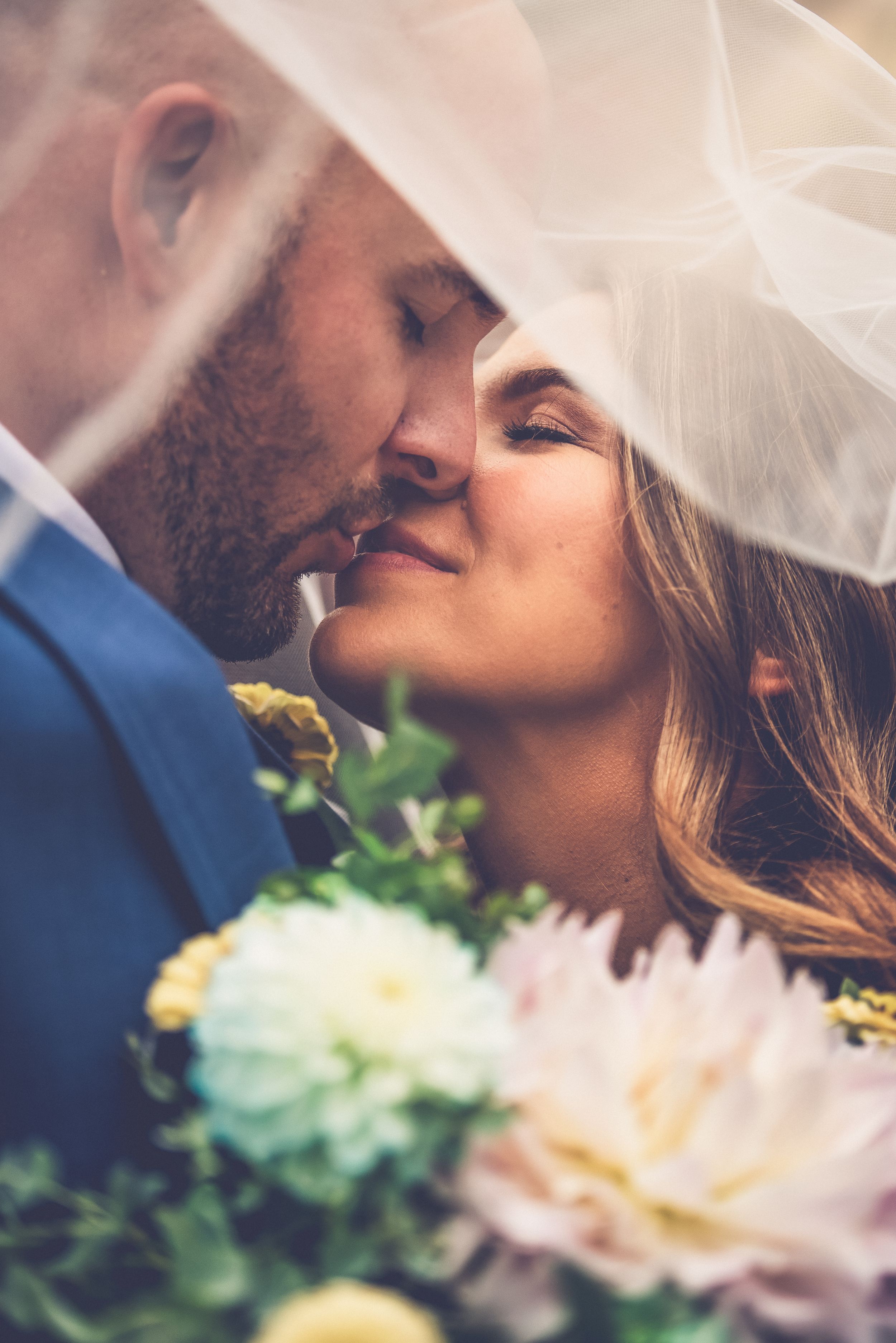 Groom kisses bride underneath veil with bouquet in view