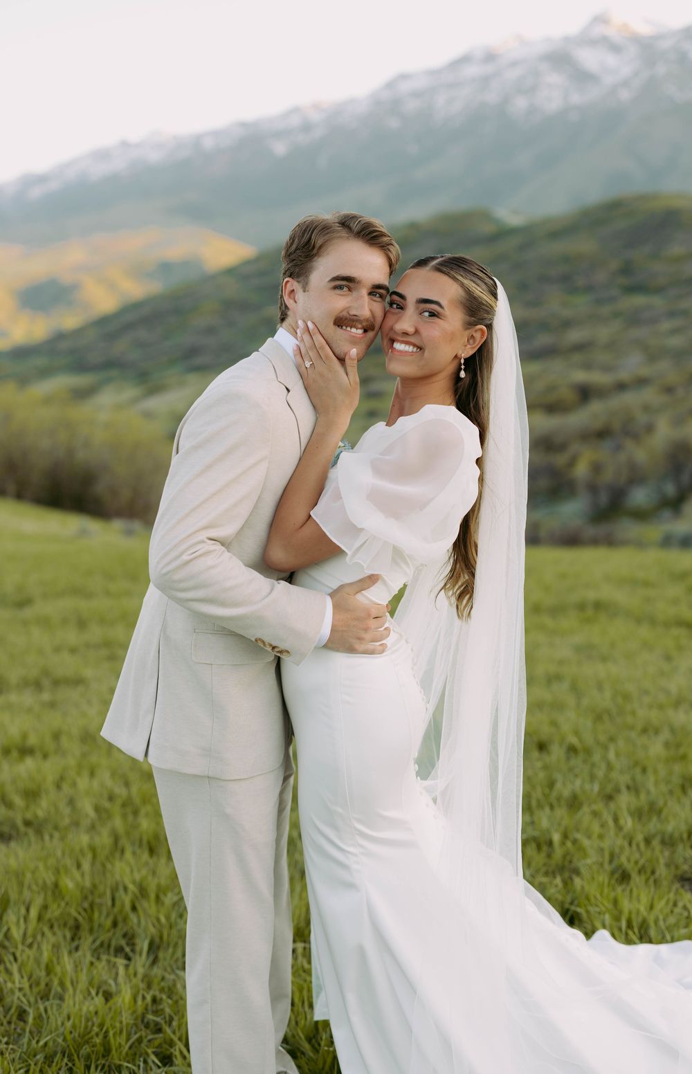 Newlywed couple in all white embrace in a mountain meadow against snow-capped peaks during sunset.