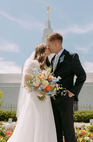 A couple shares a kiss in front of a temple spire on their wedding day, with a colorful bouquet of flowers.