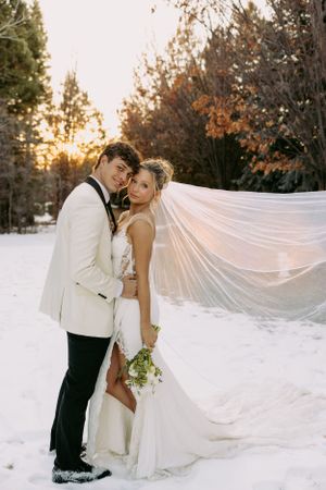 A romantic winter wedding photo with a long flowing veil catching the light at sunset in a snowy forest setting.