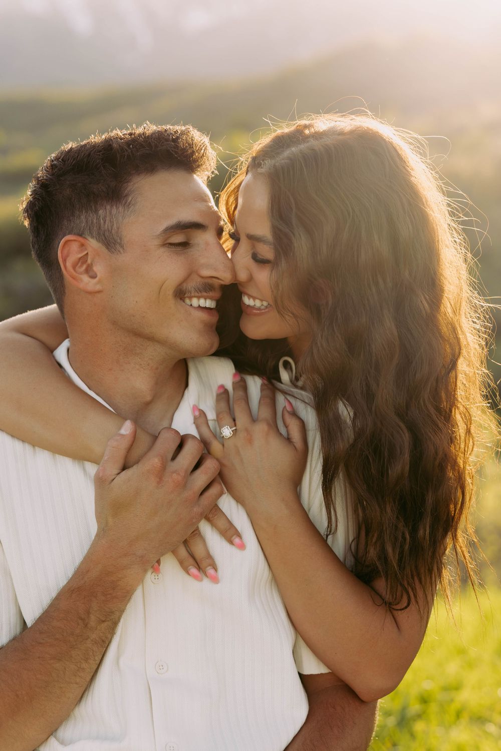 Romantic couple embraces in golden sunlight on grassy hill during sunset, captured in intimate candid moment.
