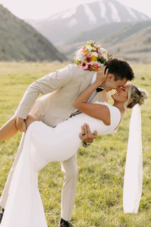 A couple in wedding attire share a romantic kiss in an outdoor mountain meadow with colorful wildflower bouquet.