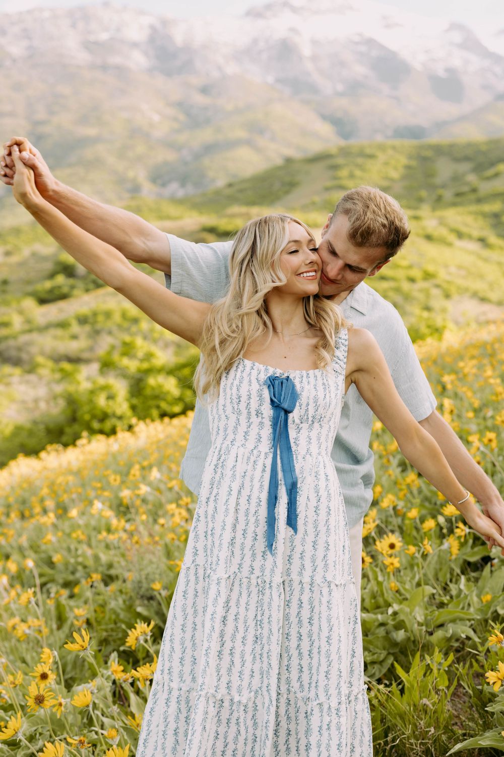 A couple embraces joyfully among yellow wildflowers in a mountain meadow on a bright summer day.
