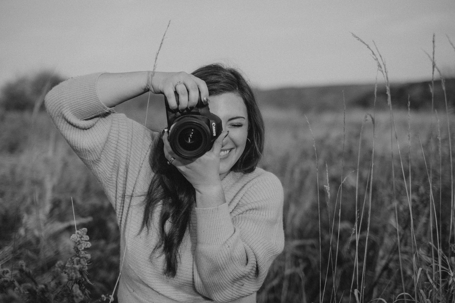 Photographer holding a Canon DSLR camera in a sunlit field wearing a white sweater and jeans.