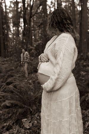A pregnant woman in a white dress and sweater stands in a forest while someone in plaid is visible in the background.