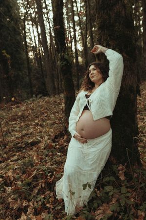 A pregnant figure in a white sweater and skirt poses gracefully against a tree trunk in an autumn forest setting.