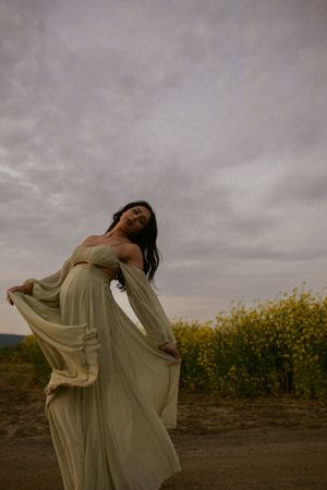 A flowing sage green dress flows in the breeze against a moody sky and yellow wildflower field at sunset.
