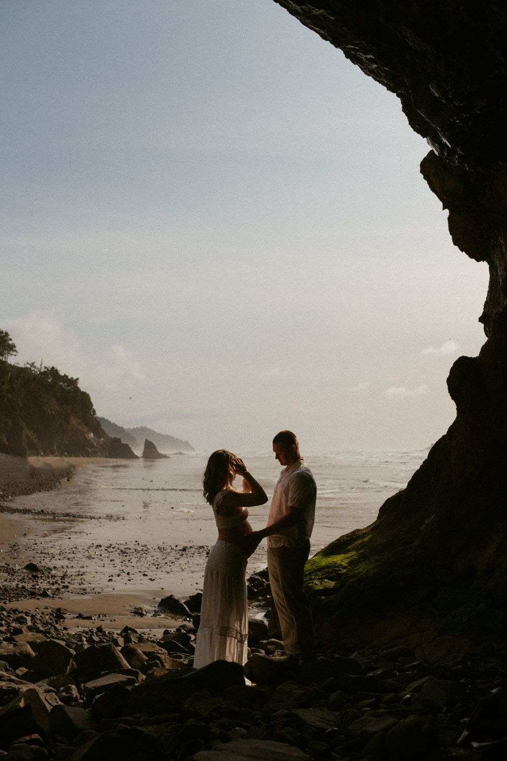 Two silhouettes share a romantic moment under a cave arch at sunset on a rocky beach with coastal cliffs in the background.