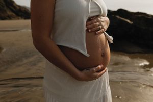 Person in white dress cradles pregnant belly on a beach at sunset, wearing beaded bracelets.