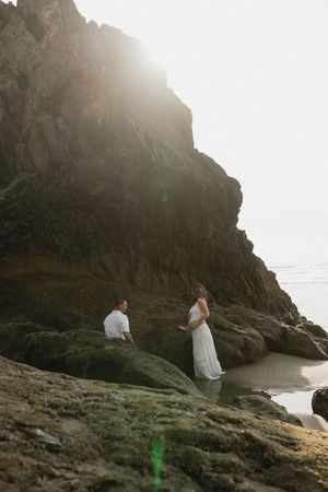 A couple sits on rocky coastal cliffs at sunset with dramatic backlighting and lens flare.