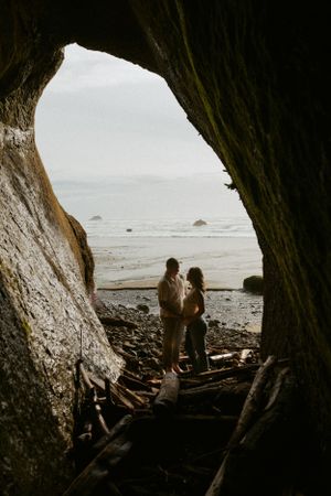 Two silhouettes stand in a cave opening overlooking a misty beach with rock formations along the Pacific coast.