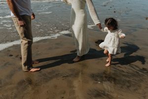 A family walks barefoot on a sandy beach together, their feet leaving footprints in the wet sand at sunset.