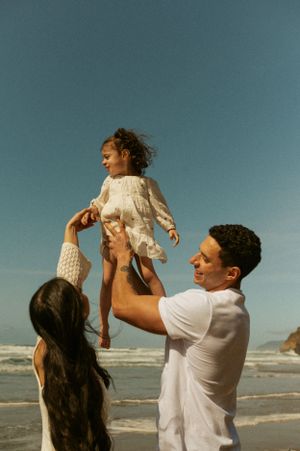Family playing and lifting child together on sunny beach with ocean waves in background.