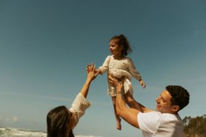 A couple lifts and plays with a young child against a blue sky at the beach during golden hour.