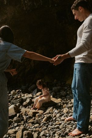 Two people holding hands while standing on rocky shore as small child plays on stones below in moody lighting.