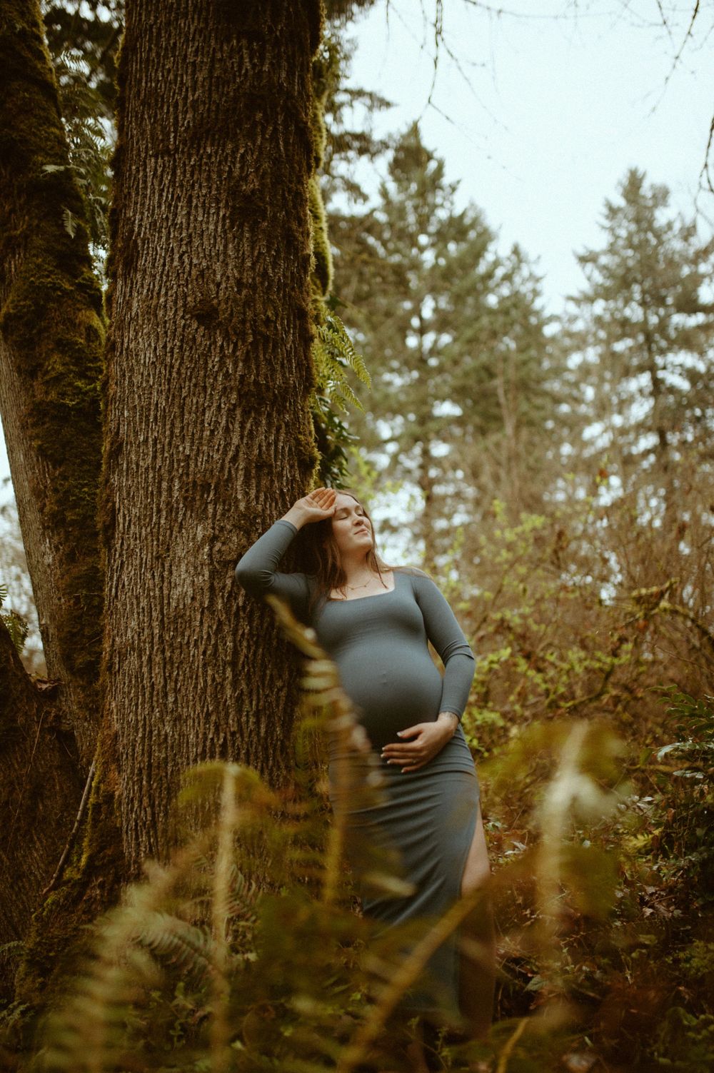 Maternity portrait of expectant mother leaning against mossy tree in forest setting.
