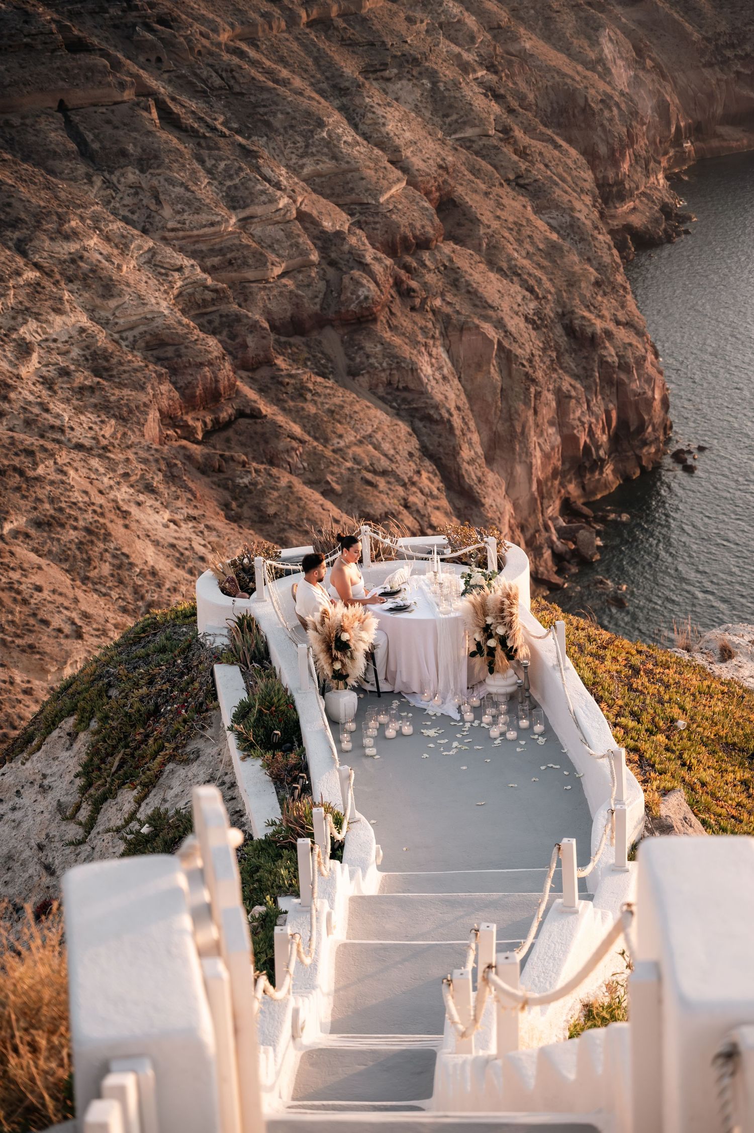 White wedding setup with floral decorations on dramatic clifftop stairway in Santorini.