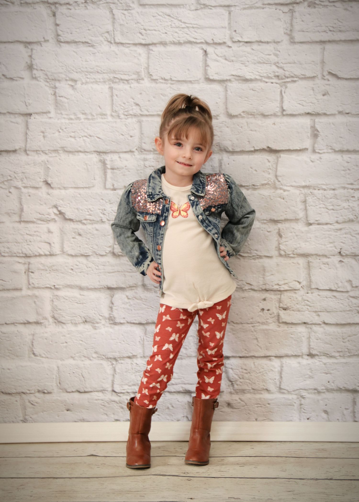 Stylish child wearing floral denim jacket, cream top, patterned leggings and brown boots against white brick wall.