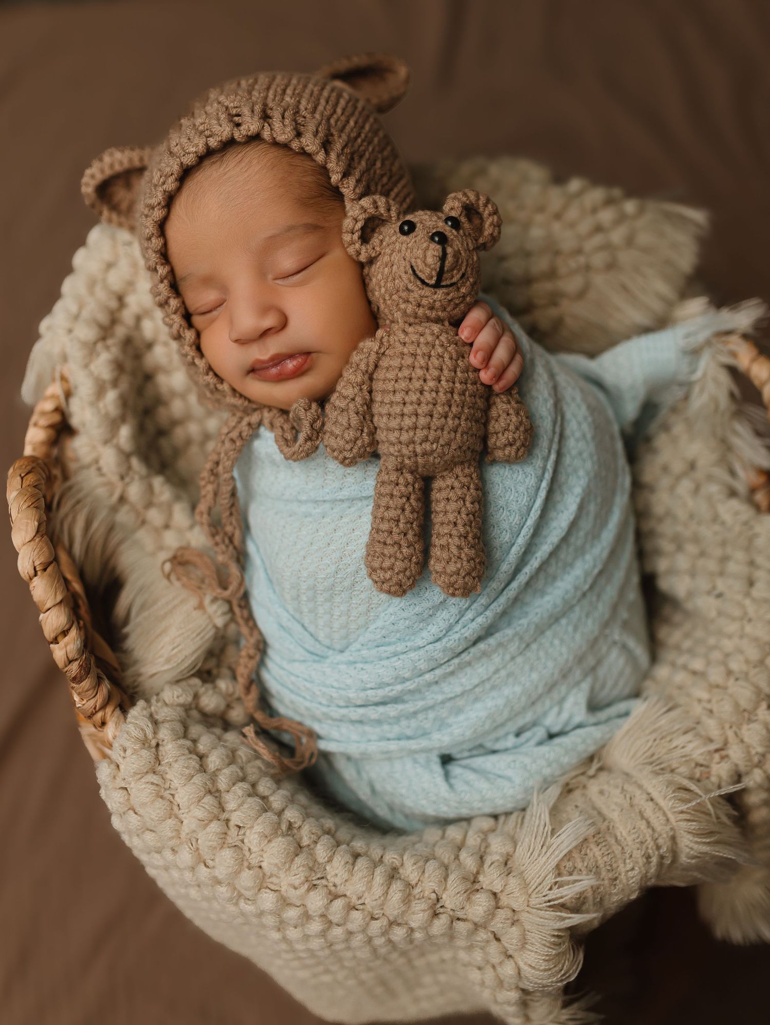 A sleeping infant swaddled in light blue fabric with a knitted hat and toy teddy bear rests in a cream-colored basket.  Colorado Springs