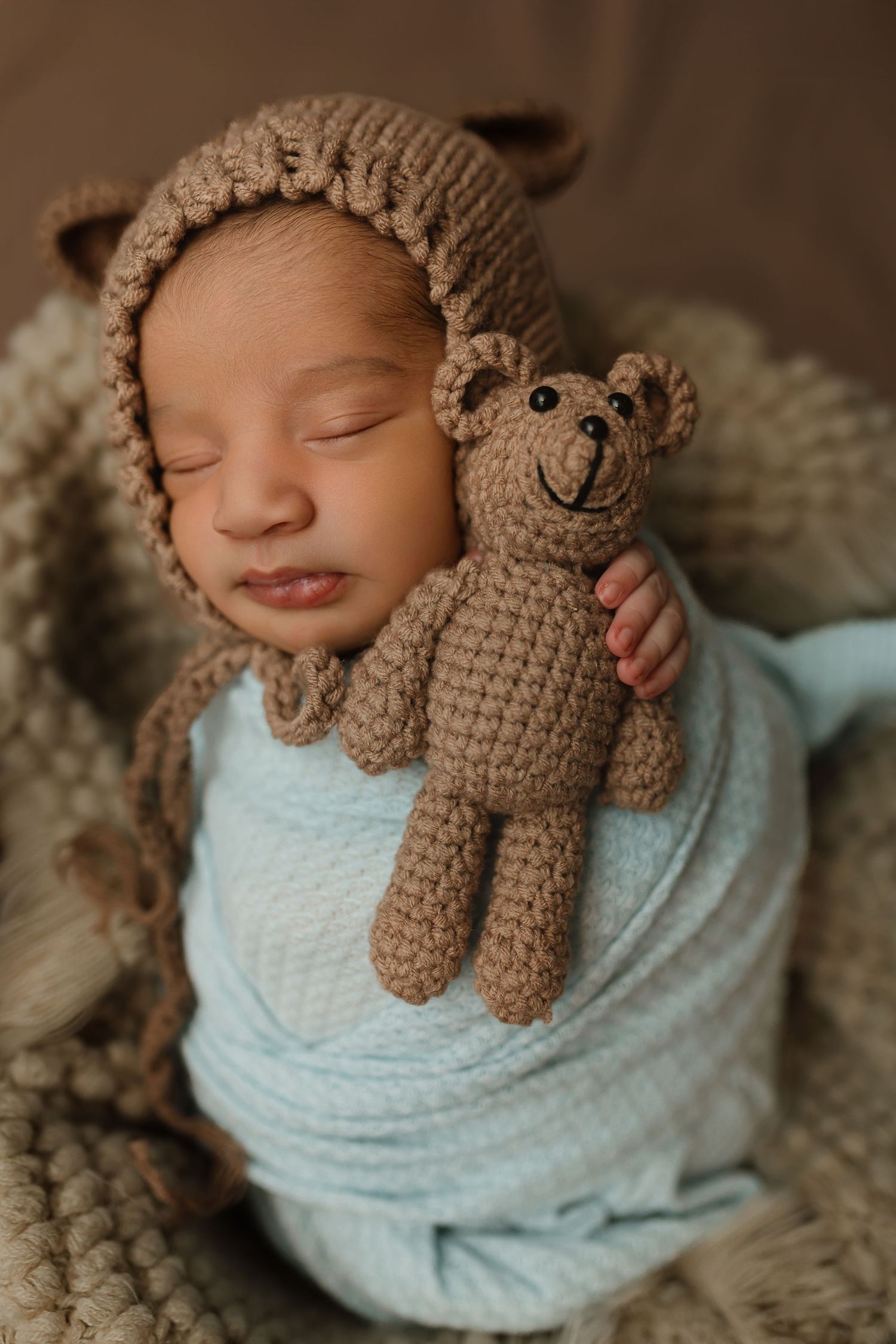 Sleeping newborn wrapped in light blue swaddle wearing brown knit bear bonnet and holding matching crochet teddy bear. Colorado Springs