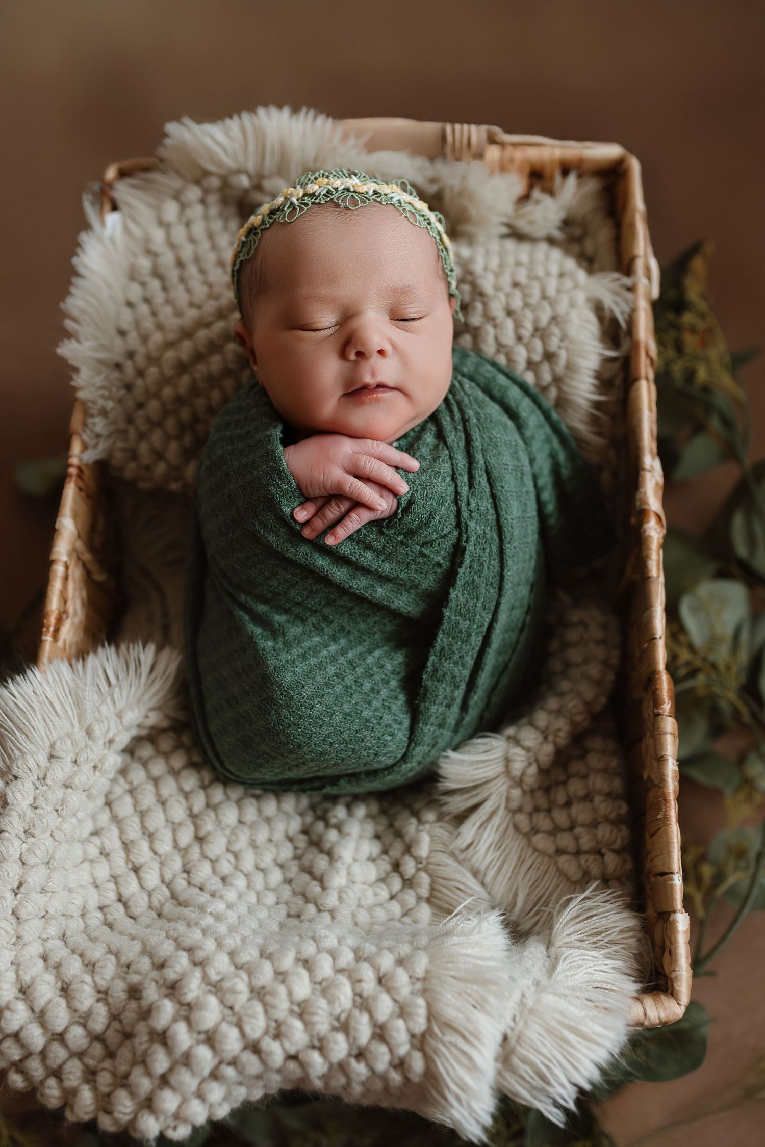 A sleeping newborn wrapped in green swaddle rests in a basket lined with cream knit blanket and greenery. Colorado Springs Photographer.