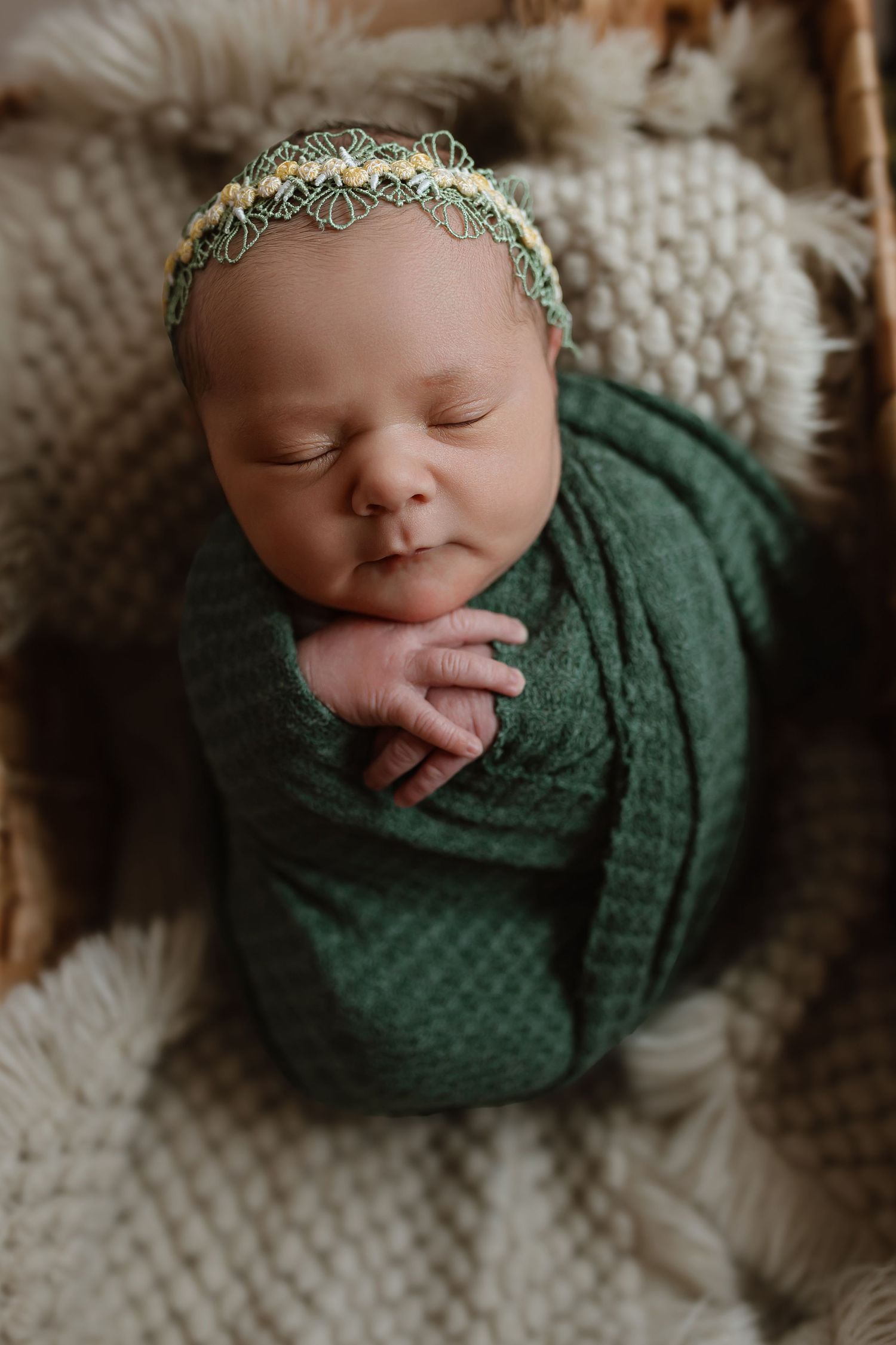 A peaceful sleeping baby swaddled in dark green fabric with a delicate floral headband rests on a cozy cream blanket. Colorado Springs Photo