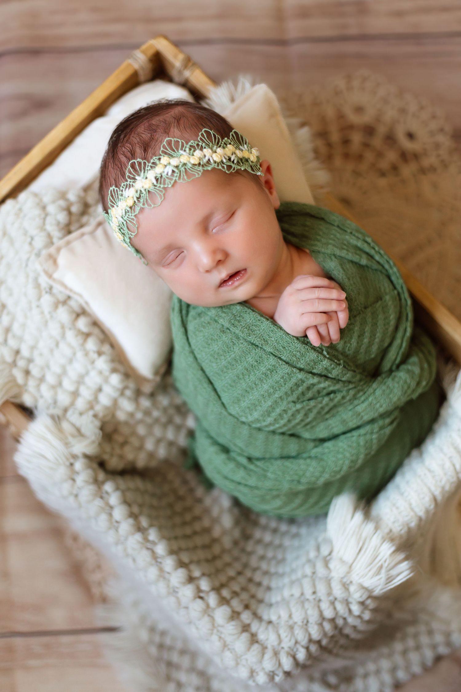 Sleeping newborn swaddled in soft green wrap on cream knitted blanket with delicate gold headband.