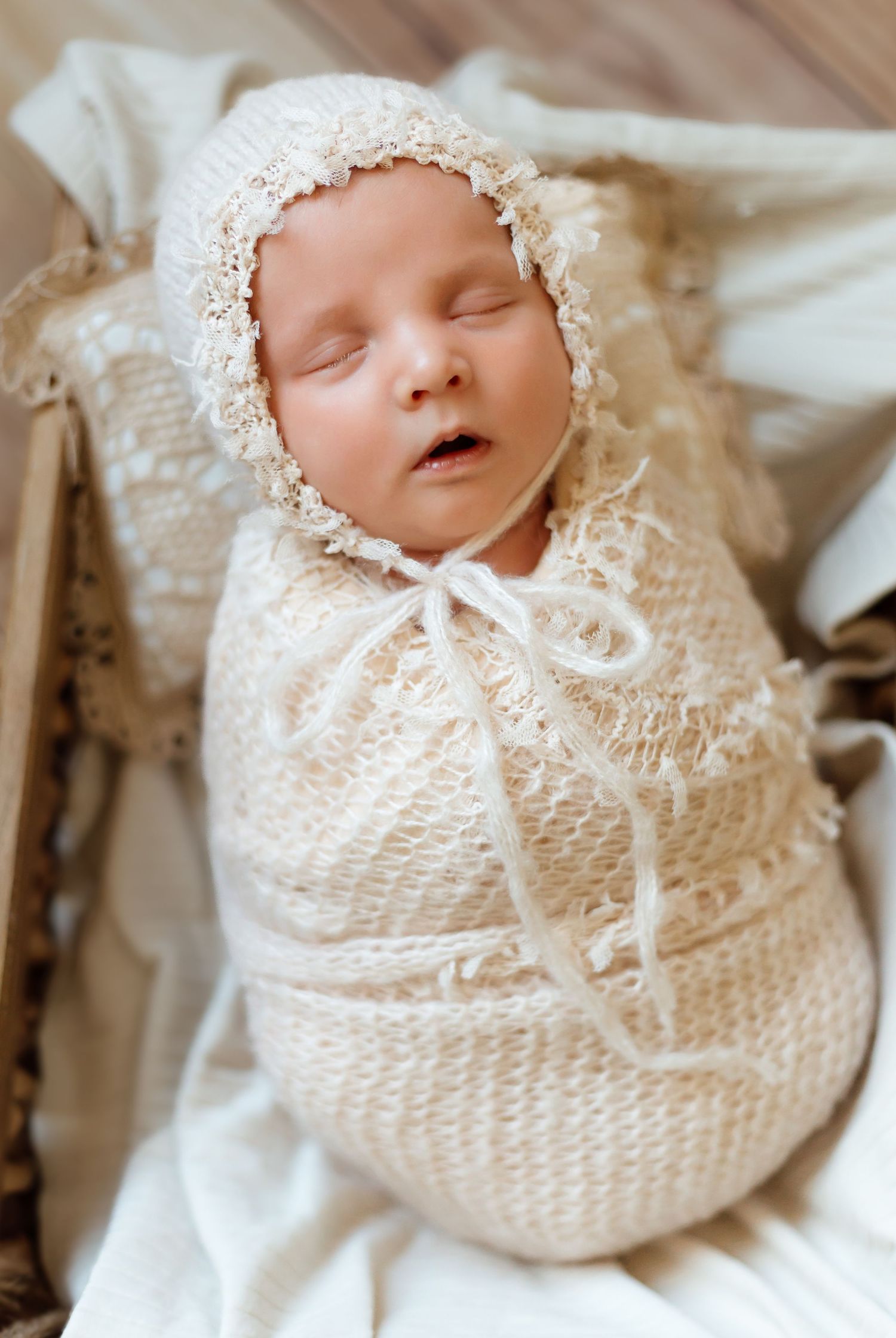 A sleeping newborn swaddled in delicate cream-colored knit fabric with lace details against white bedding.