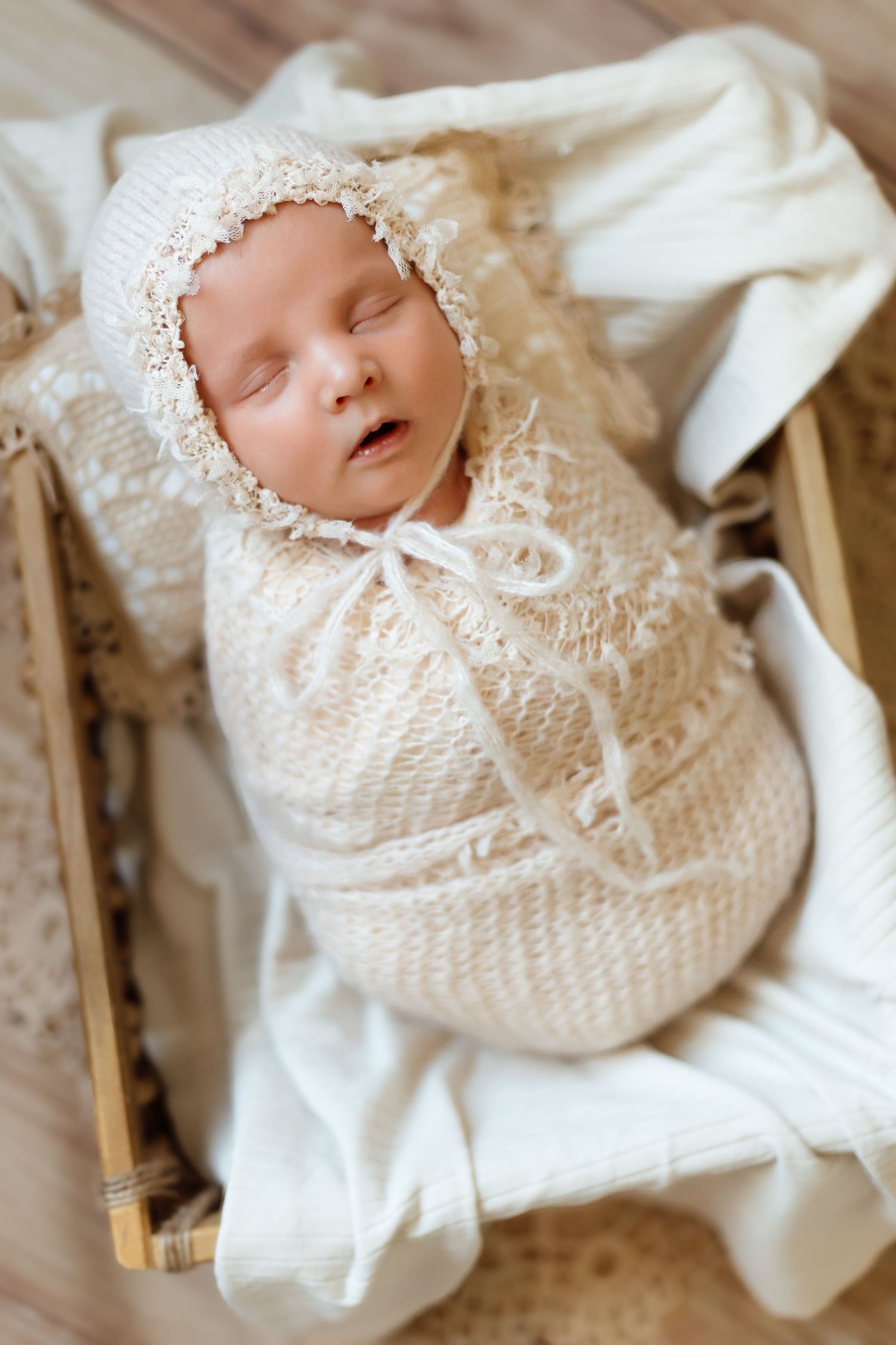A peaceful newborn baby wearing a delicate white knitted bonnet and christening gown lies on soft fabric.