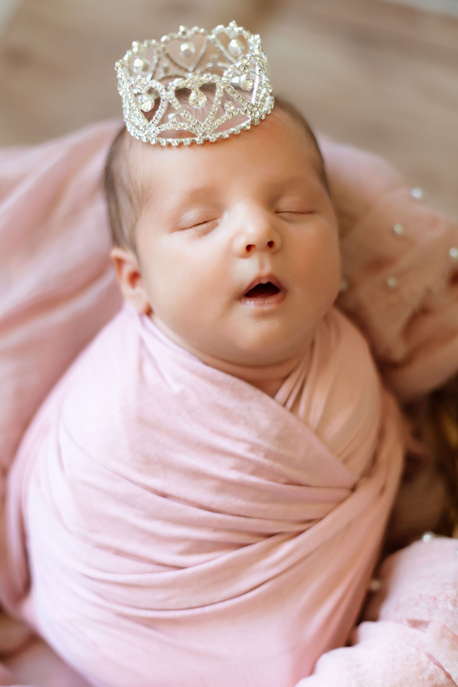 A sleeping newborn baby wearing a delicate crystal crown tiara wrapped in soft pink fabric against neutral bedding.