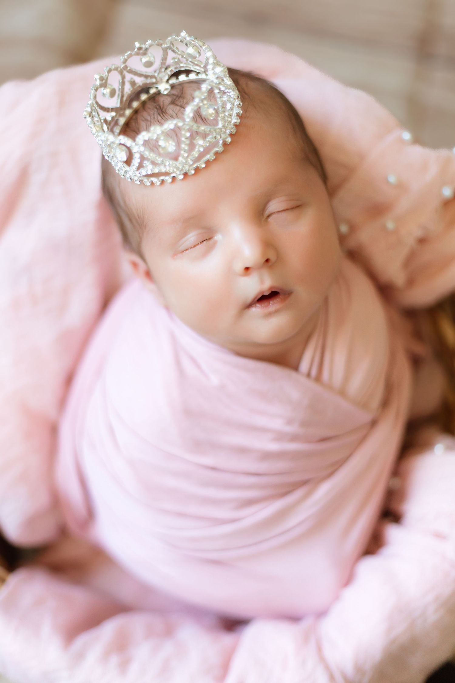 Sleeping newborn wrapped in soft pink fabric wearing delicate silver crown tiara against blurred background.