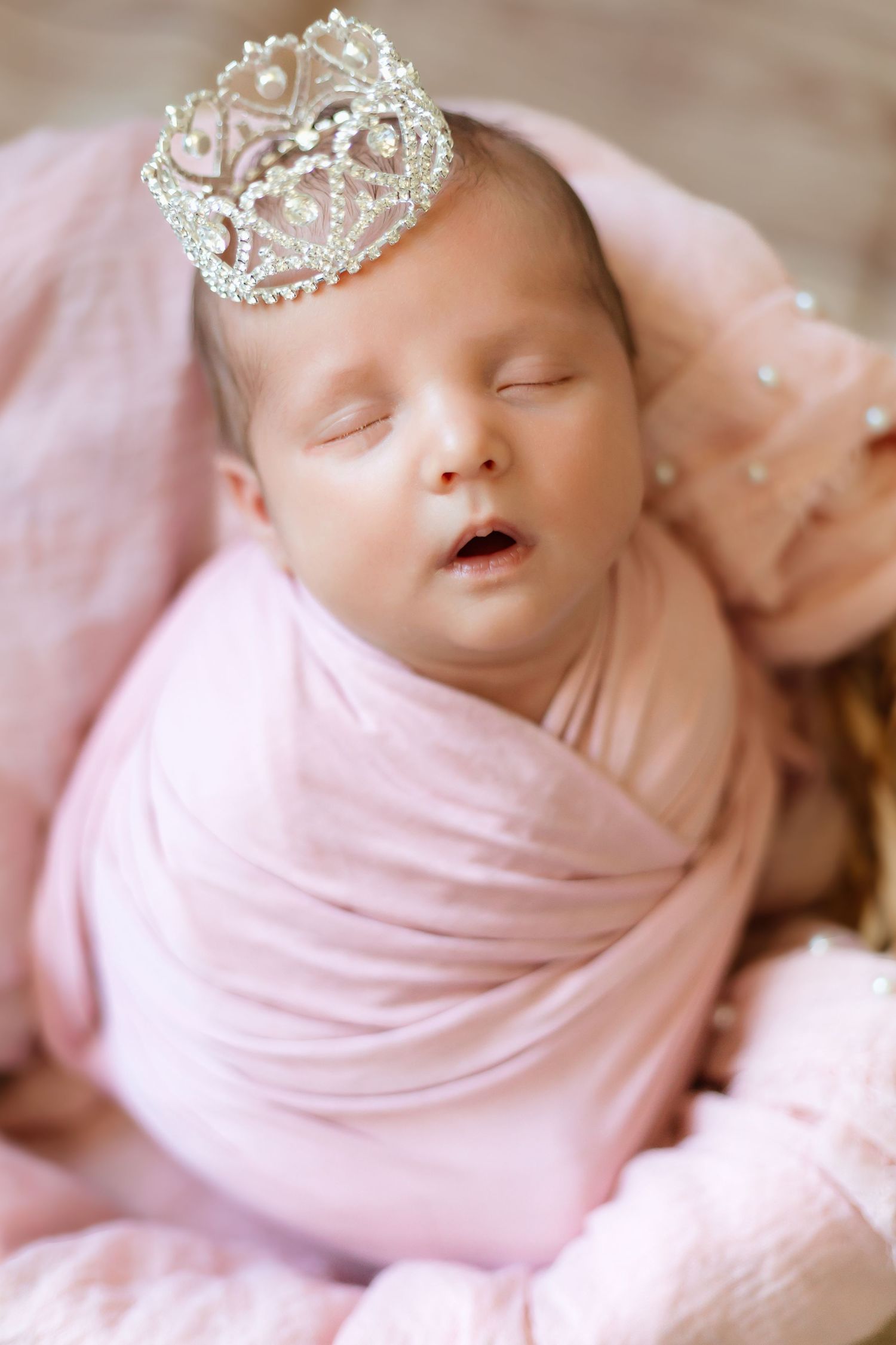 A sleeping newborn baby wrapped in soft pink fabric wearing a delicate crystal tiara on their head.
