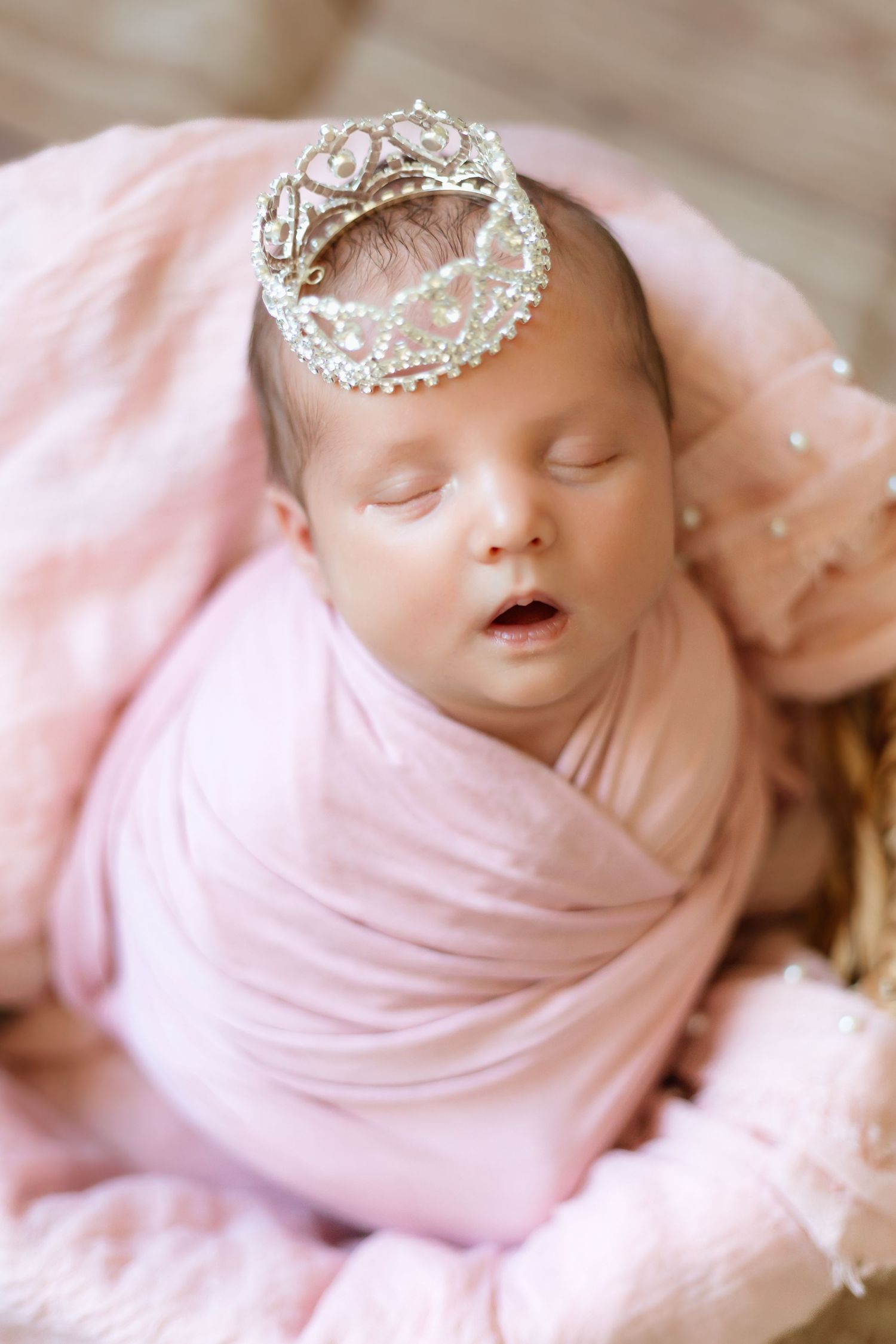 Sleeping newborn baby swaddled in soft pink fabric wearing a delicate crystal crown accessory.