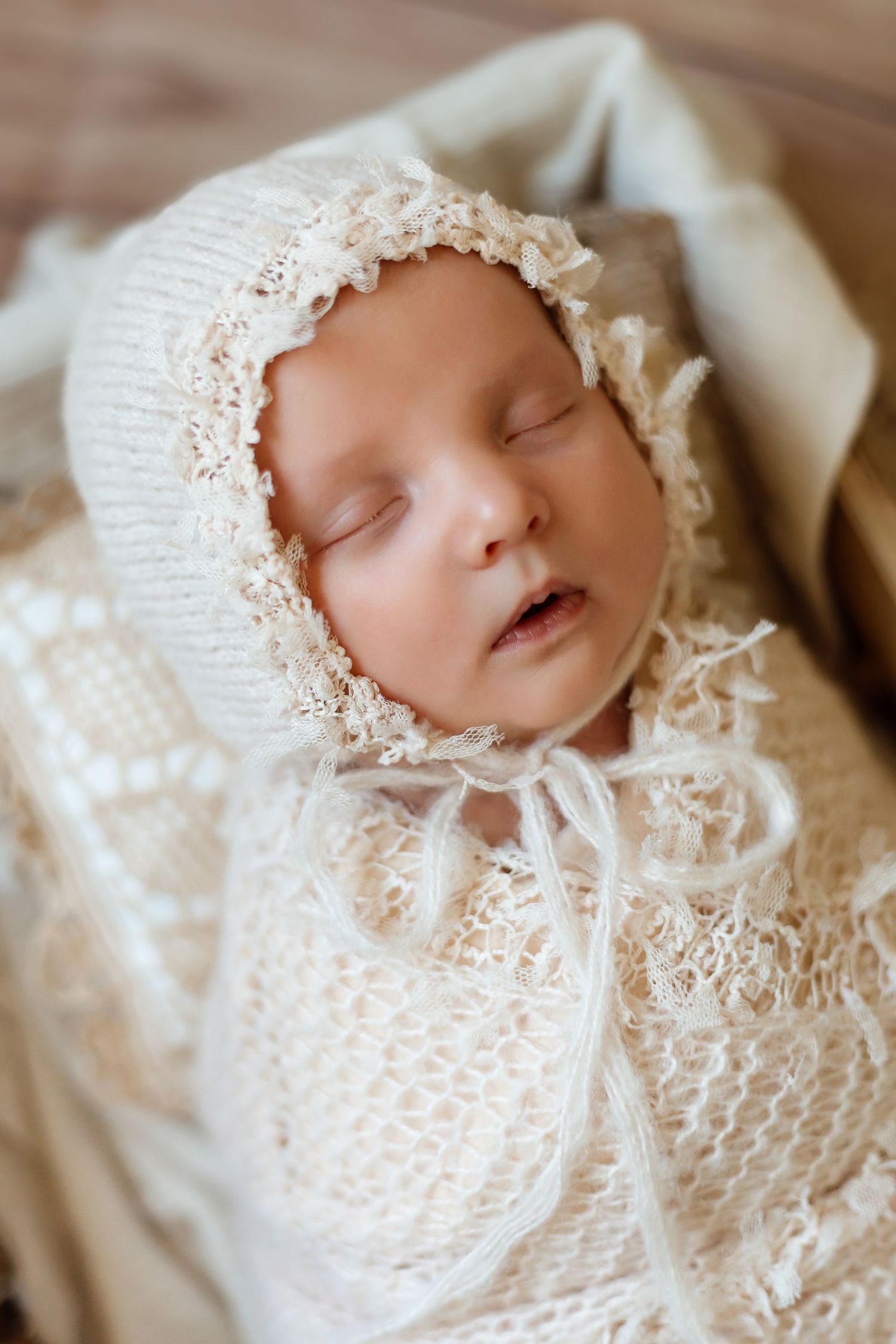 Sleeping newborn wearing delicate white lace bonnet and dress on cream colored blanket.