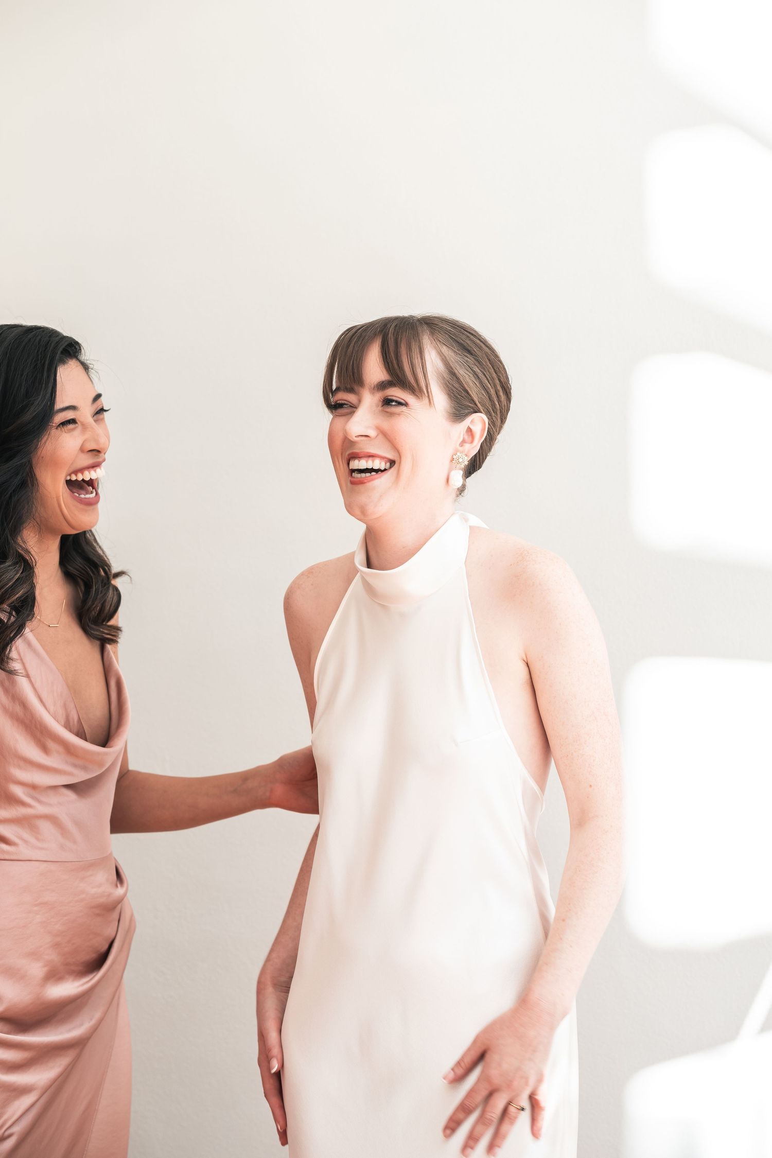 Two people in elegant dresses share a joyful moment against a white background.