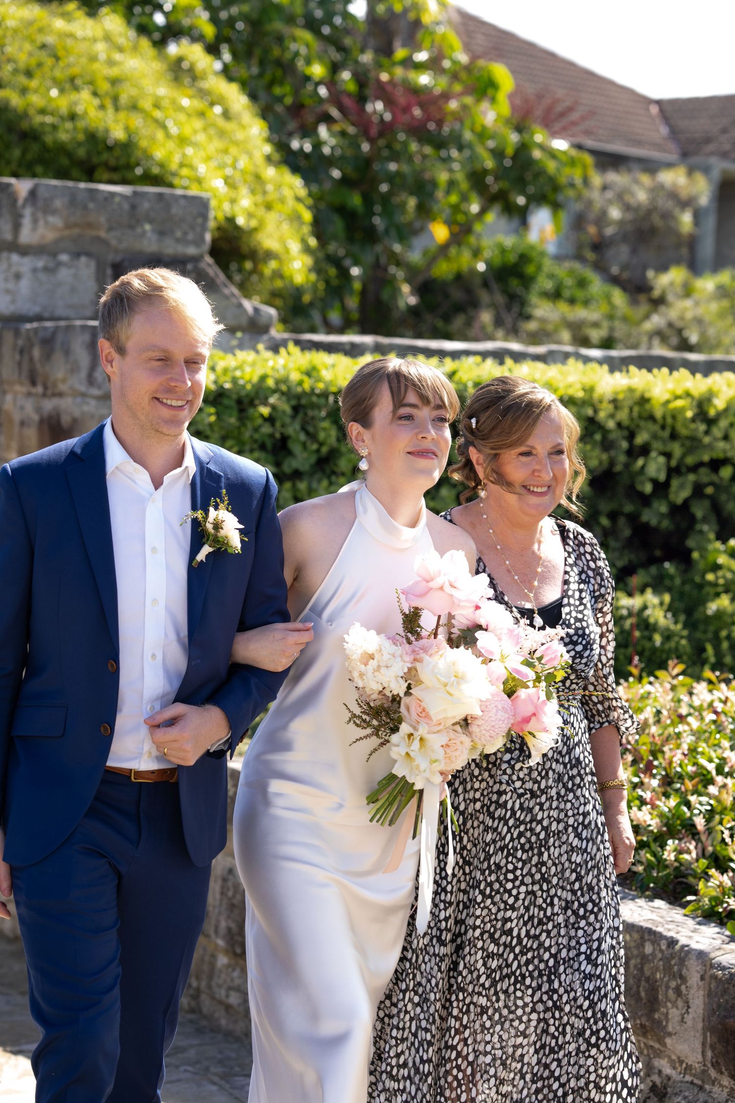 A sunny outdoor wedding photo with three people walking together in formal attire.