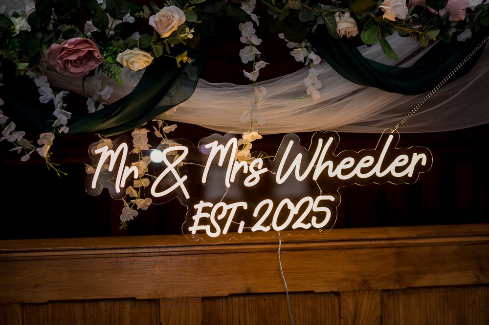 Custom neon wedding sign glows against wooden backdrop decorated with roses and white fabric.