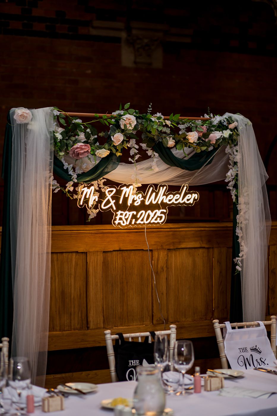 Elegant wedding reception decor features illuminated name sign with floral garland and white draping.
